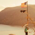 A hiker stands with an American flag overlooking a picturesque lake and mountains during fall.