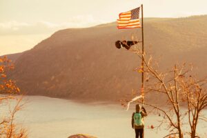 A hiker stands with an American flag overlooking a picturesque lake and mountains during fall.